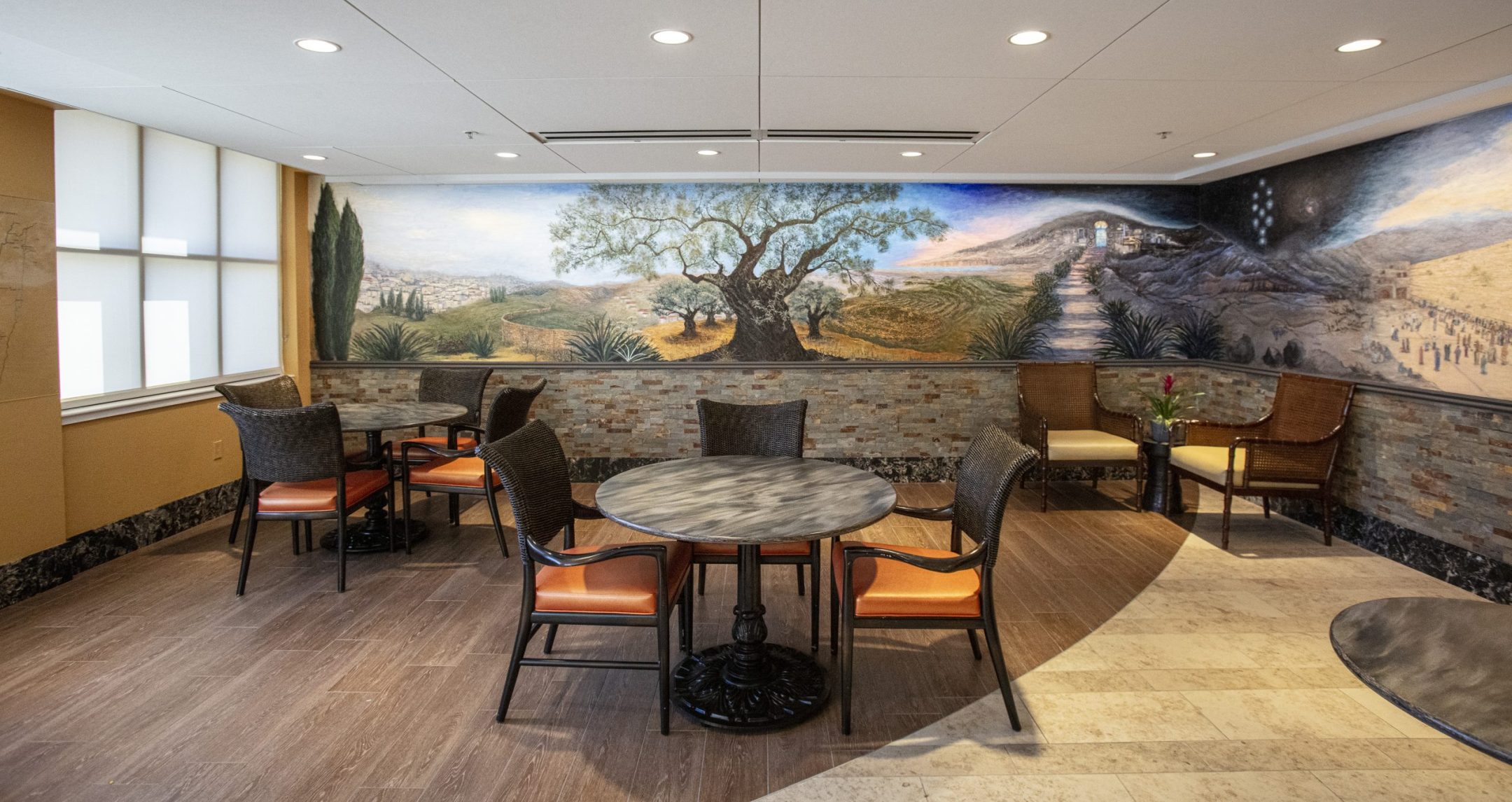 A small seating area with round tables and wicker chairs sits by a mural of a tree, landscape, and pathway. Warm lighting and a modern, cozy atmosphere invite you to relax—or schedule a tour to experience it in person.