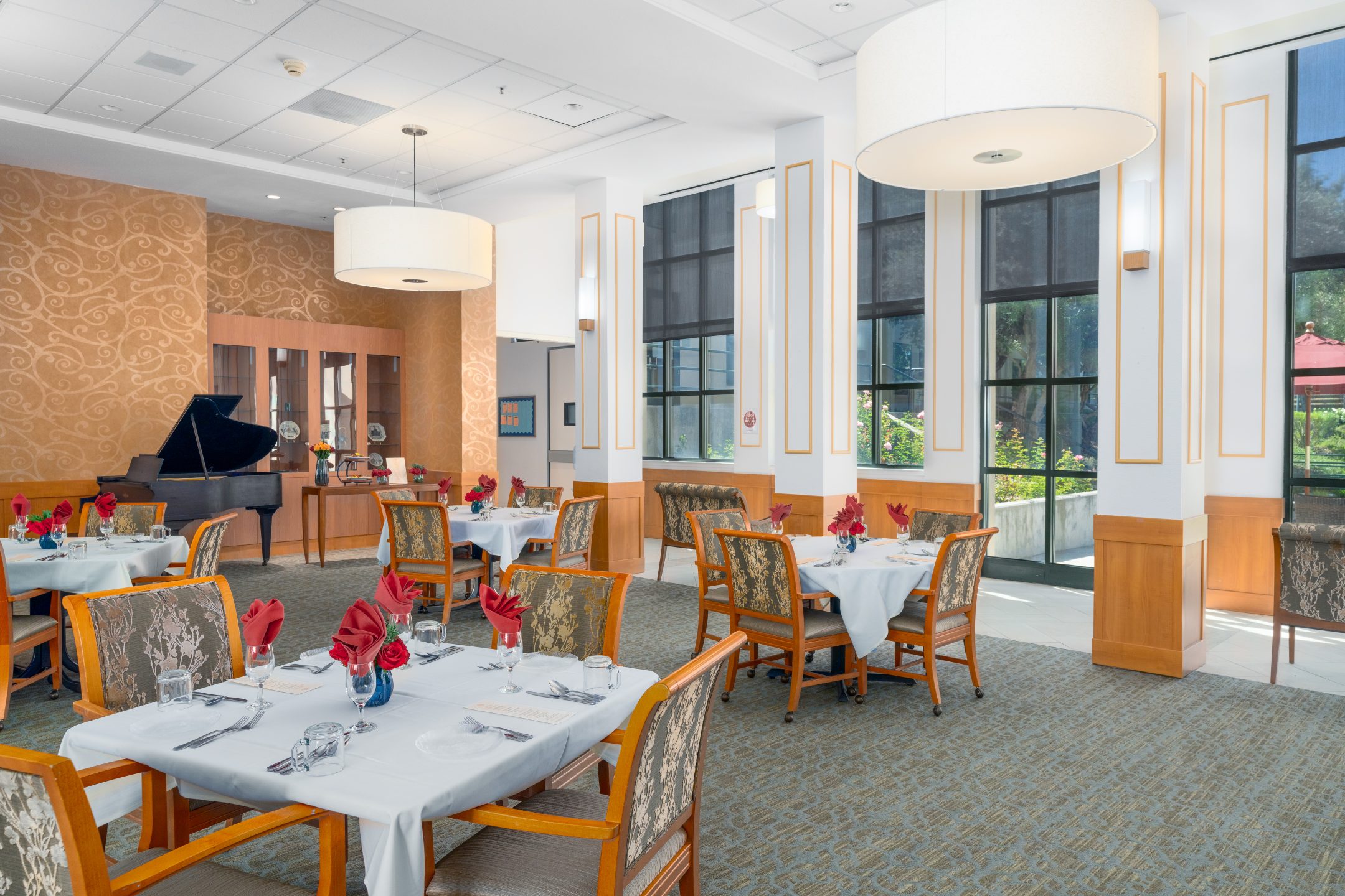 Bright, elegant dining room with tables set for a meal, wooden chairs, large windows, chandeliers, and a grand piano in the background. Each table has white linens and red napkins folded decoratively for an unforgettable culinary experience.