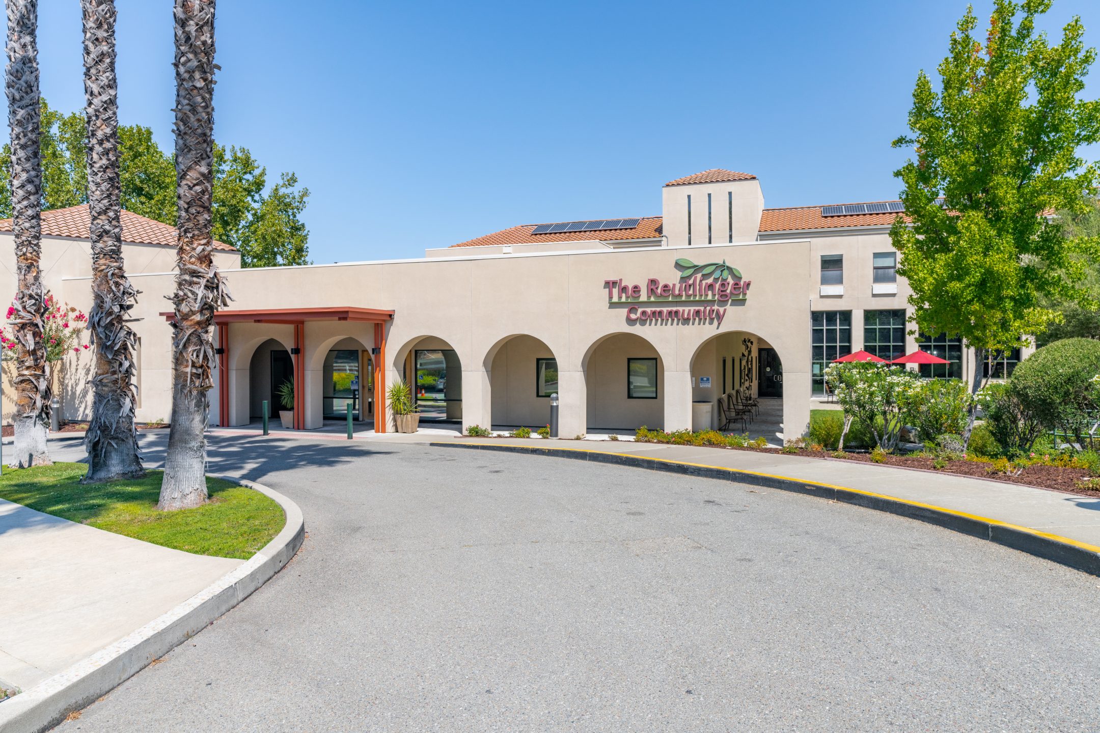 A beige stucco building with arched entryways, “The Reutlinger Community” sign, palm trees, and solar panels on the roof sits on a sunny day with a curved driveway in front—perfect for your next brochure or to download for more information.