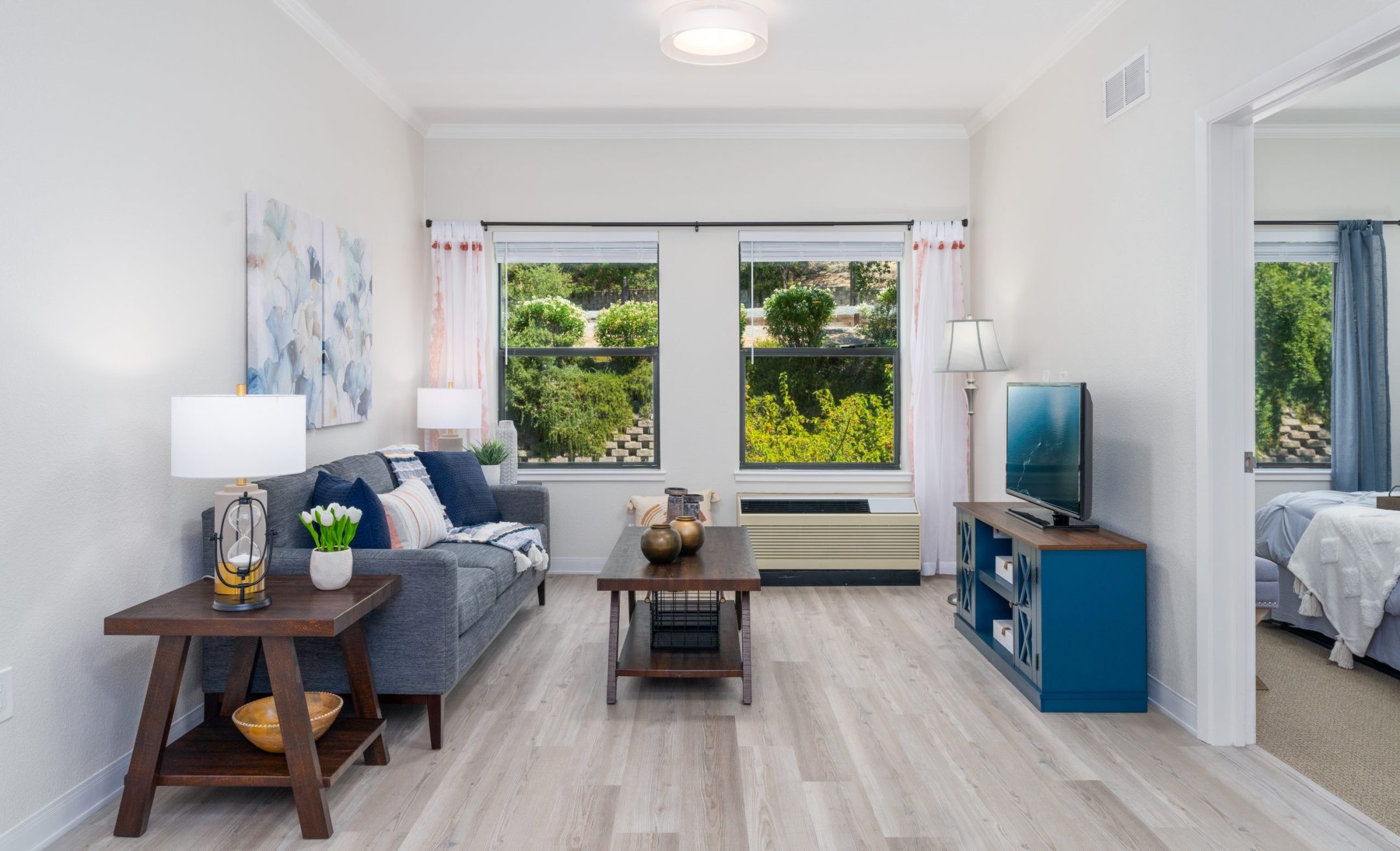 Bright living room with light wood floors, gray sofa, coffee table, side tables, and a TV on a blue stand. Large windows reveal garden views and natural light. A bedroom is visible through an open doorway, showcasing versatile living options.