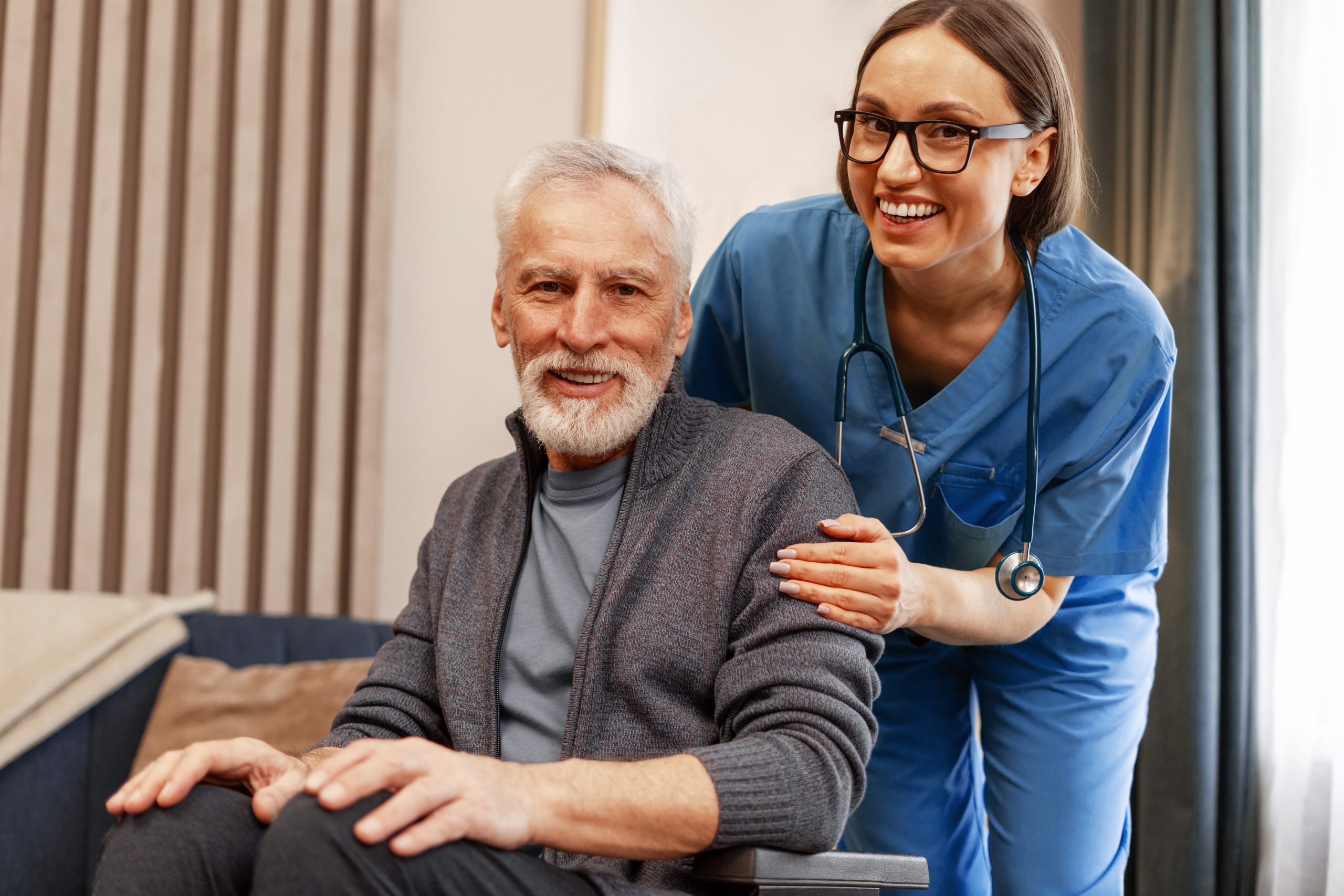 A smiling older man sits in a wheelchair while a cheerful female healthcare worker in blue scrubs and glasses stands beside him, gently holding his arm in a supportive manner during his rehabilitation.