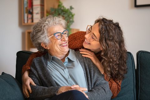 Older woman and younger woman with curly hair, both wearing glasses, sit close together on a couch, smiling and embracing joyfully in a cozy living room—an example of the warmth found in The Reutlinger living options.