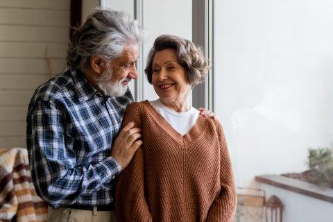 An elderly couple stands by a window, looking at each other and smiling warmly. The man gently rests his hand on the womans shoulder. They appear happy and comfortable together in a cozy indoor setting.