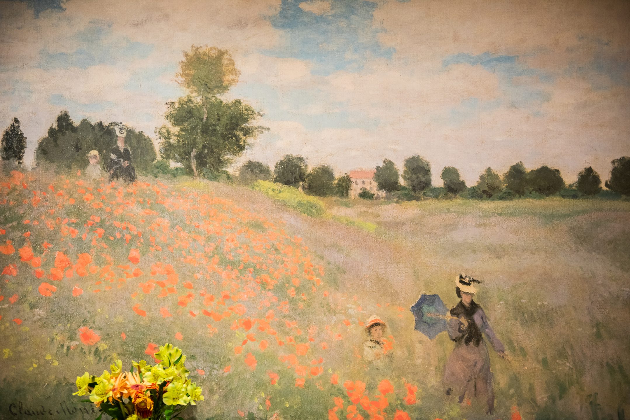 A painting of a sunny field with red poppies, green grass, and scattered trees evokes peace—much like the comfort found in skilled nursing care. Two women, one holding a blue umbrella, and a child stroll along the hillside under a partly cloudy sky.