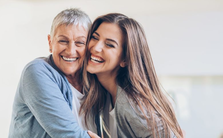 Two women, one older with short gray hair and one younger with long brown hair, are smiling and hugging each other closely, showing happiness and affection.