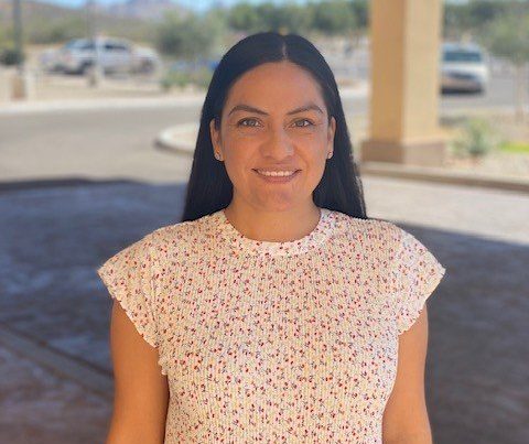 A woman with long dark hair, wearing a white short-sleeve blouse with a pink and purple floral pattern, stands outdoors and smiles at the camera near The Reutlinger senior dining area, with parked cars and trees in the background.